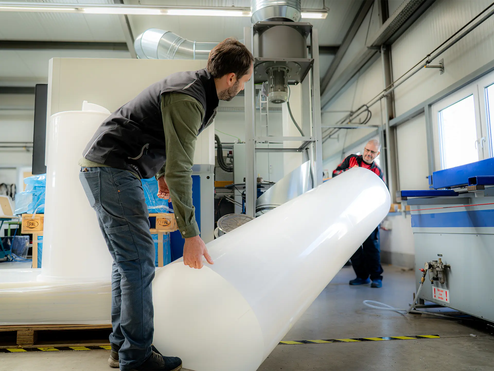 Zwei Arbeiter in einer Industriehalle heben und positionieren ein großes glänzendes weißes Kunststoffrohr