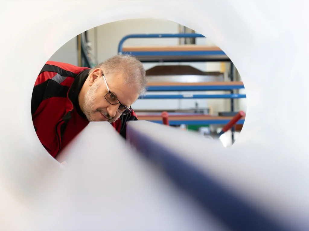 Mann mit Brille und roter Arbeitsjacke beugt sich durch eine runde Öffnung und inspiziert die Walzen einer Fertigungsmaschine