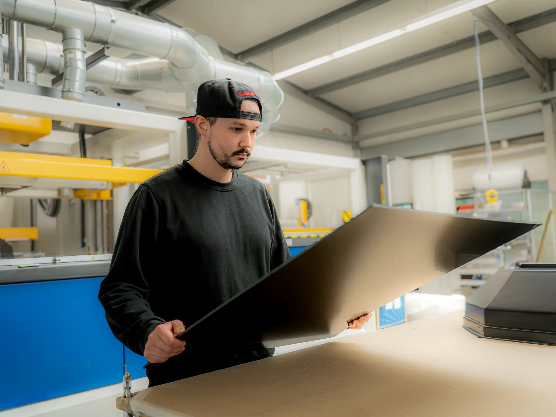 Mann in Werkstatt mit Rückwärts-Baseballcap und Gehörschutz prüft großes schwarzes Blech an einer Arbeitsstation