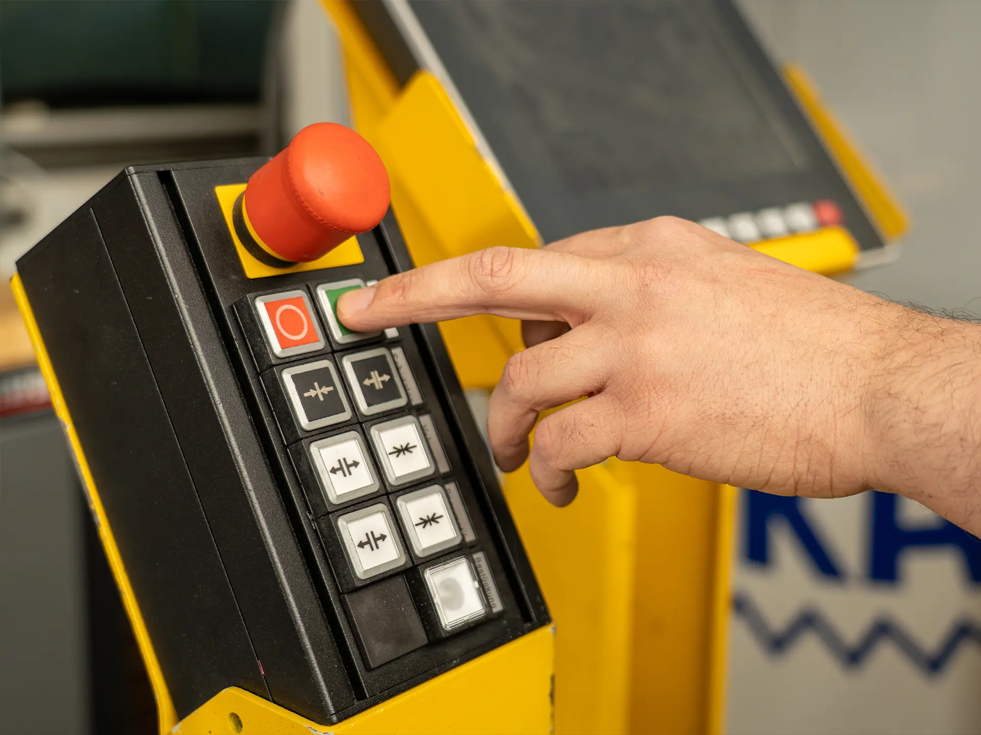 Hand drückt grünen Startknopf an gelbem Industrie-Bedienfeld neben großem roten Notausschalter