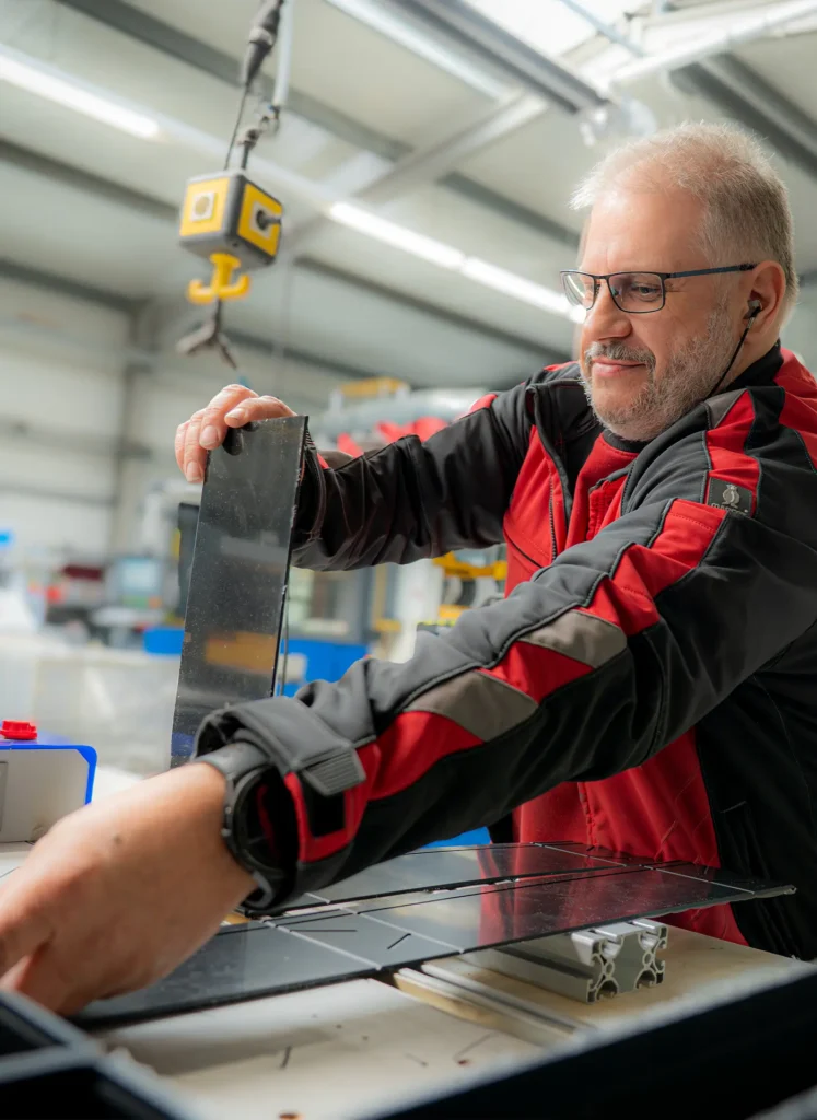 Handwerker mit Brille und rotschwarzer Arbeitsjacke richtet ein schwarzes Blech auf einer Werkbank in einer Industriehalle aus
