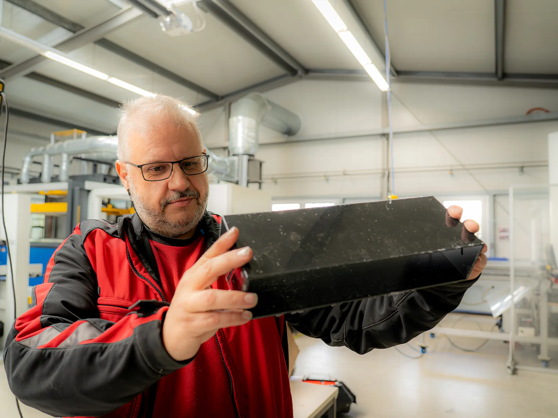 Älterer Mann mit Brille in rotschwarzer Arbeitsjacke begutachtet in einer Werkhalle ein schwarzes rechteckiges Bauteil