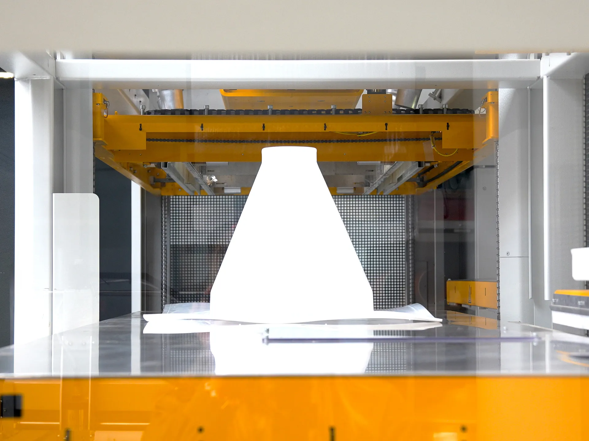 Industrielle Fertigungsmaschine mit gelbem Portalarm über einer Bauplatte, auf der ein großes weißes, konisch geformtes Bauteil steht