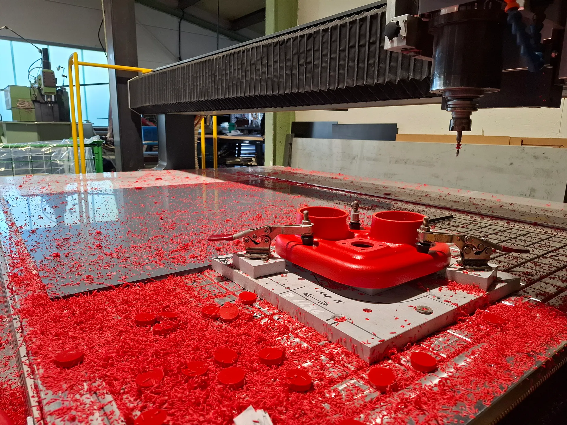 CNC‑Fräsmaschine bearbeitet ein rotes Kunststoffteil; Arbeitstisch und Boden mit roten Spänen bedeckt, Werkstück mit Klammern fixiert