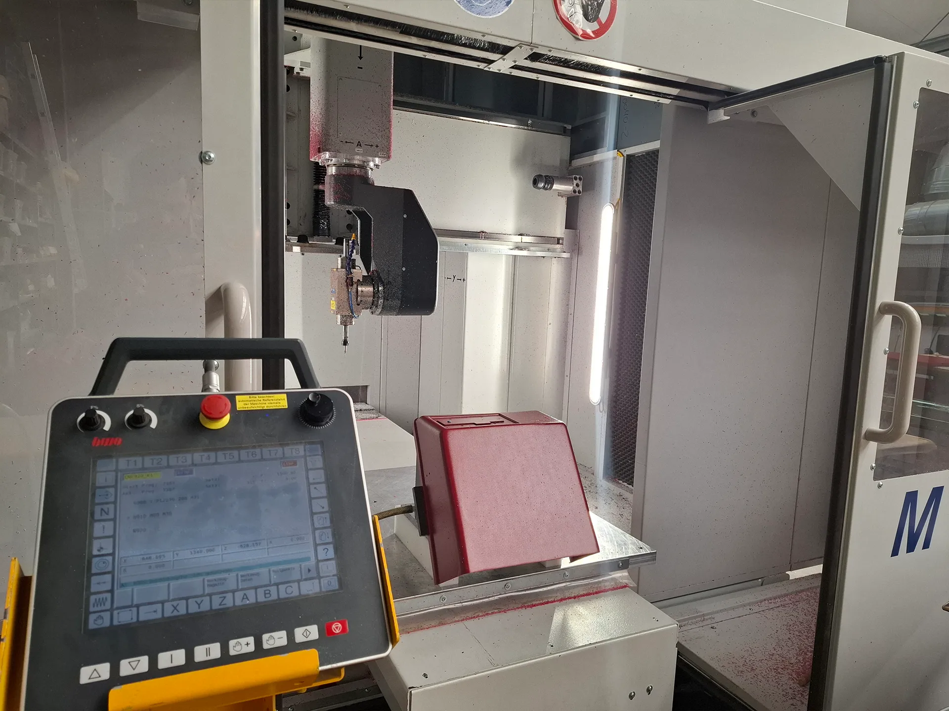 CNC-Bearbeitungsmaschine: Spindelkopf über rotem Werkstückhalter im geschlossenen Gehäuse, Bedienpult mit Not-Aus im Vordergrund