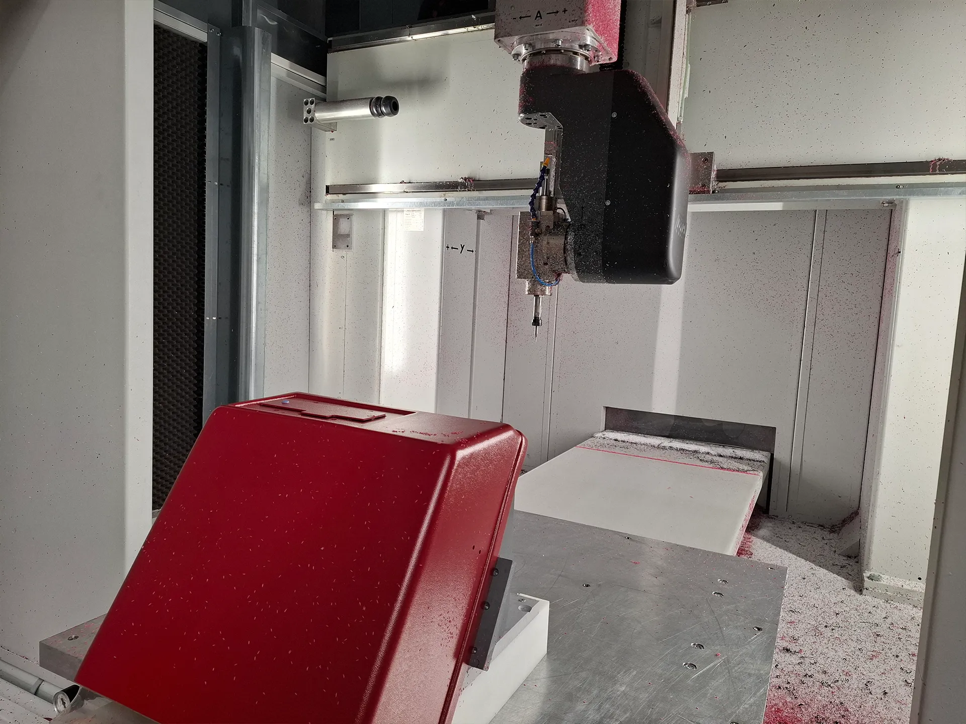 Innenraum einer CNC‑Fräsmaschine: schwarze Spindel über weißem Werkzeugtisch, rote Schutzhaube und rosa Kunststoffspäne