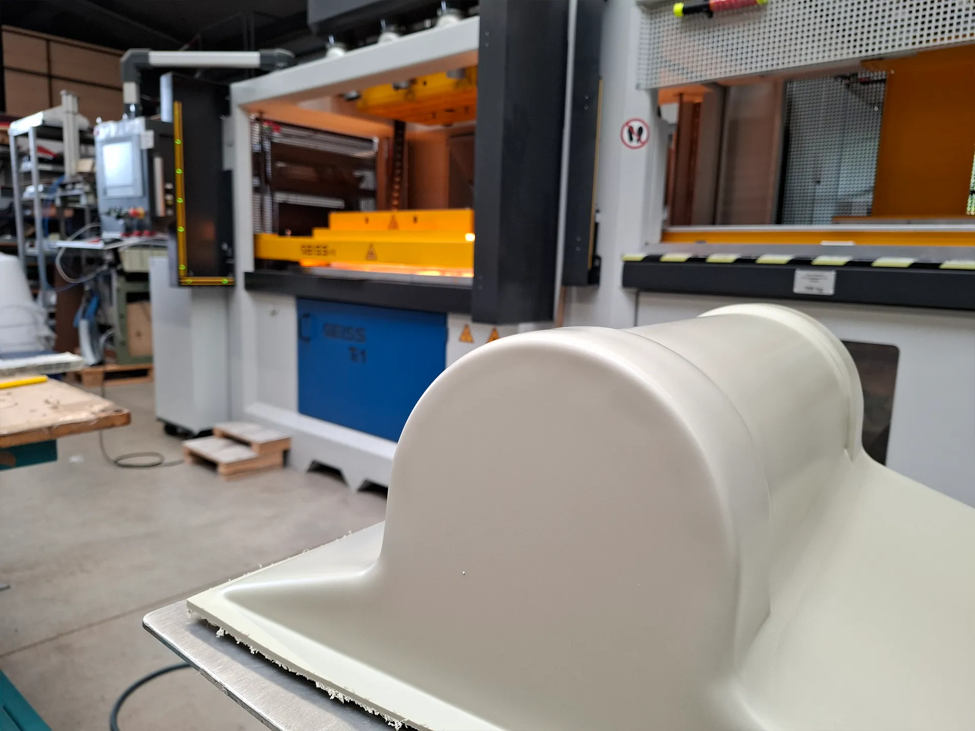 Geformtes beiges Kunststoffteil im Vordergrund vor einer gelb-orangen Industriemaschine mit Bedienfeld in einer Werkstatt