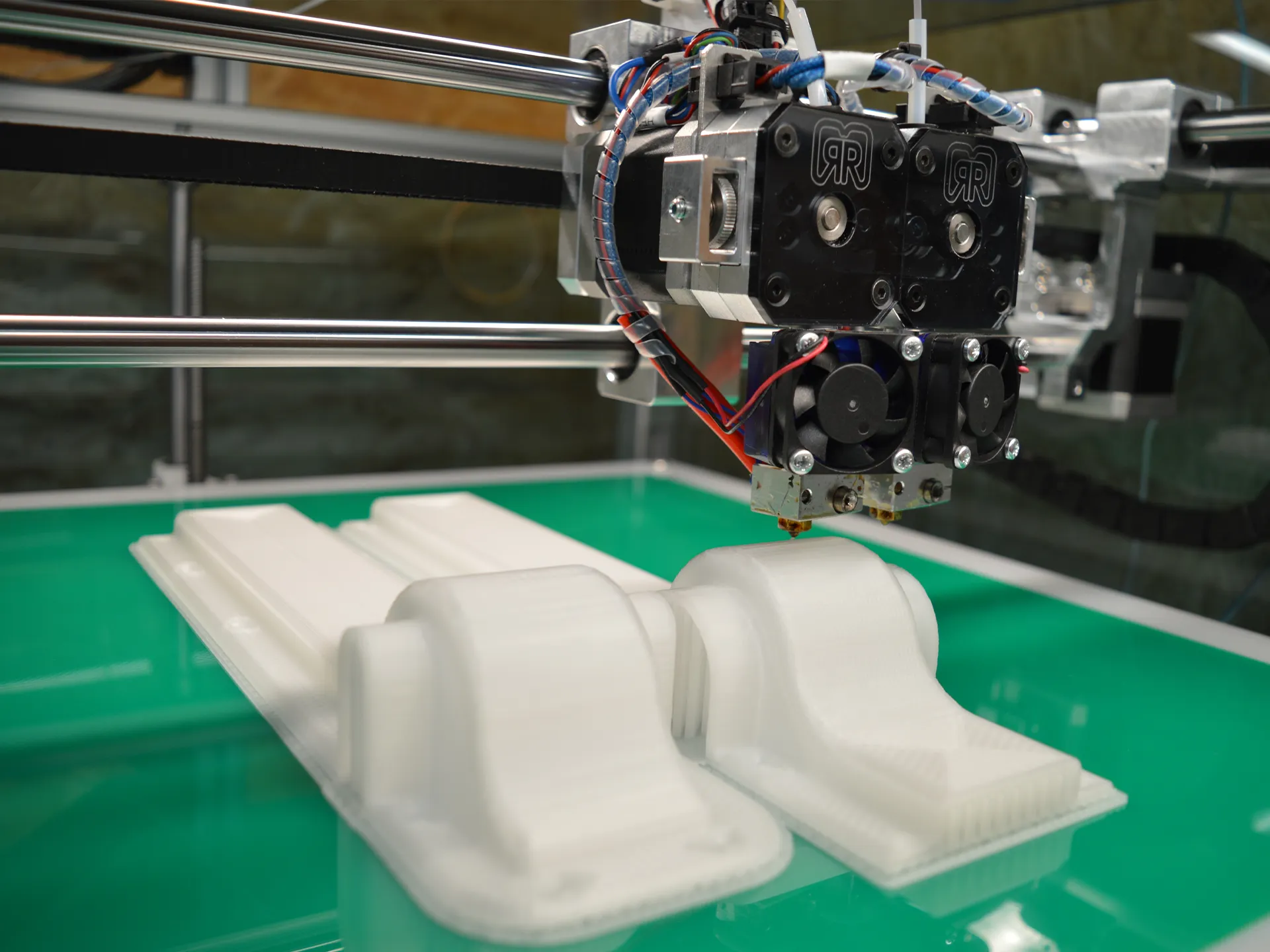 Doppel-Extruder einer 3D-Drucker-Montage mit Lüftern und Kabeln druckt weiße Kunststoffteile auf grünem Druckbett
